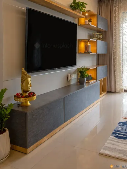 The TV unit in this living room combines dark grey laminate with gold-accented open shelves. A Buddha statue adds a touch of serenity to the entertainment area.