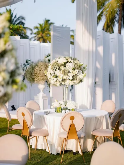 A pristine white table setting for an outdoor wedding. The large, spherical arrangement of white flowers serves as a stunning centerpiece, perfectly complementing the elegant chairs and the natural beauty of the surroundings.