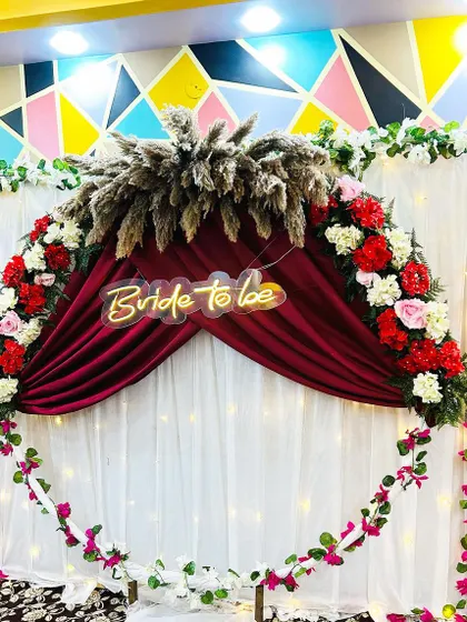 A close-up of the 'Bride to be' backdrop, showing the details of the floral arch, the red velvet drape, and the glowing neon sign.