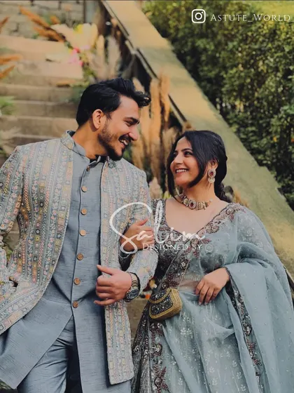 A happy couple during their celebrations. I designed the groom's outfit, a modern Indo-Western ensemble featuring a grey-blue kurta and a patterned open jacket, perfect for a sangeet or engagement ceremony.