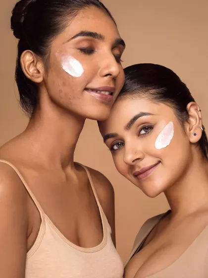 This image captures a moment of quiet connection between two models, both with dabs of sunscreen on their cheeks. The soft lighting and gentle interaction highlight the product's smooth texture while promoting a message of shared skincare and diverse beauty.