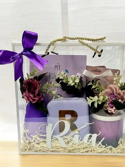A chic and personalized Rakhi gift for a sister named Ria. This purple-themed hamper, presented in a clear box, includes a custom initial, a mini travel case, a candle, and other thoughtful items.