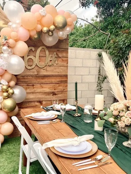 An elegant outdoor setup for a 60th birthday celebration. The rustic wooden table, green runner, and pastel balloon arch create a sophisticated and warm garden party vibe.