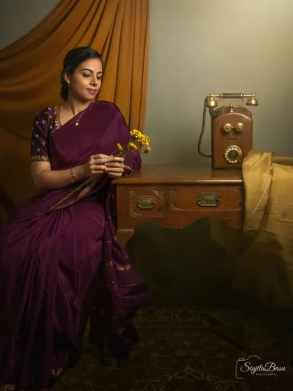 A vintage-inspired portrait featuring a woman in a beautiful purple saree, seated next to an antique telephone. The props and warm lighting create a nostalgic scene, telling a story of waiting and quiet contemplation.
