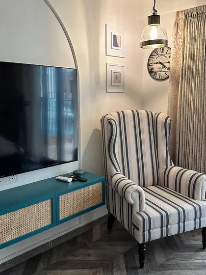 A cozy reading nook featuring a classic striped wingback chair and a teal TV console with cane-fronted doors. The herringbone wooden flooring adds another layer of texture.