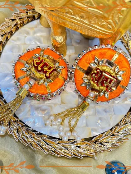 These orange "Ram Ram" hangings are set on a beautiful mother-of-pearl tray. The golden tassels and intricate embroidery make them a luxurious addition to your Rakhi decor.