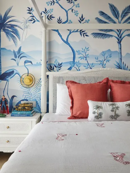 This boy's room features a stunning blue and white tropical landscape wallpaper. The classic white four-poster bed and rattan nightstand create a timeless look, while pops of red in the bedding add energy.