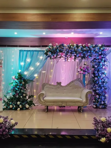 This is an example of an elegant stage I designed for a wedding reception. The setup features a beautiful floral arch with purple and white flowers over a classic white lounge sofa, with soft curtain drapes and fairy lights in the background.