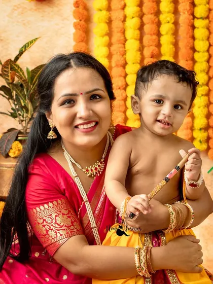 A classic portrait of a mother holding her baby dressed as Krishna, both smiling for the camera.