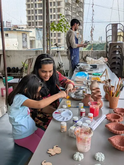A mother and daughter share a creative moment, painting their pottery pieces together on our rooftop terrace. You can see a variety of unpainted diyas and pots ready for color.