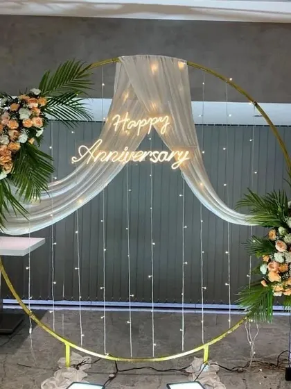 This elegant circular backdrop is perfect for anniversaries or engagements. The golden ring is draped with sheer white fabric and fairy lights, accented with two beautiful floral arrangements and a 'Happy Anniversary' neon sign.