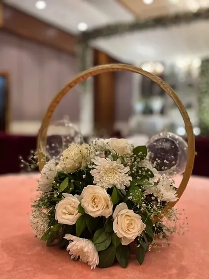 A close-up of a delicate and modern table centerpiece. This arrangement at Crowne Plaza Okhla features white roses and baby's breath inside a golden ring structure with hanging glass votives.