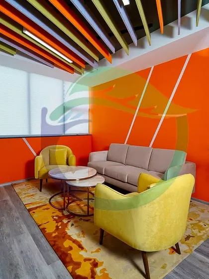 This lounge area features large planters with Areca Palms and other foliage plants. The greenery softens the bold orange wall and complements the yellow and beige furniture, creating a balanced and inviting space for relaxation.