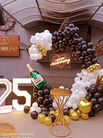 A stylish 25th anniversary or birthday party setup in a restaurant. The decor features a black, white, and gold balloon ring, a champagne foil balloon, and light-up '25' numbers.