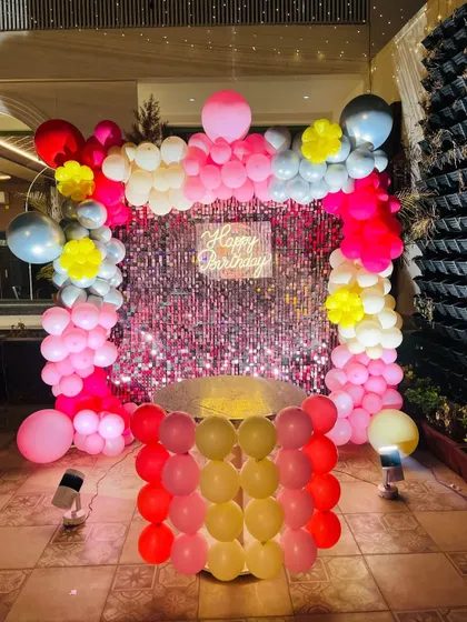 A glamorous indoor party backdrop featuring a pink shimmer wall. The wall is framed by a balloon arch in shades of pink, red, and white, with a "Happy Birthday" neon sign.
