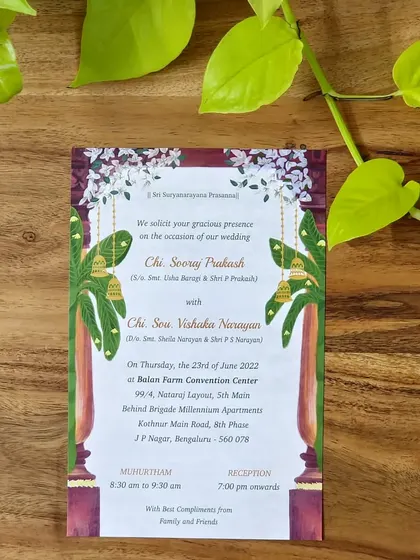 A photo of the final printed wedding invitation, showcasing a traditional South Indian theme. The design features an arch of jasmine flowers, banana leaf plants on either side of classic pillars, and hanging brass bells, all beautifully printed for a tangible keepsake.