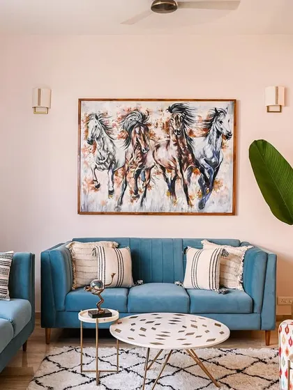 This living area is a playful mix of colors and patterns, with cool blue sofas, a quirky patterned armchair, and a large painting of horses. A tall plant and large window connect the space to the greenery outside.