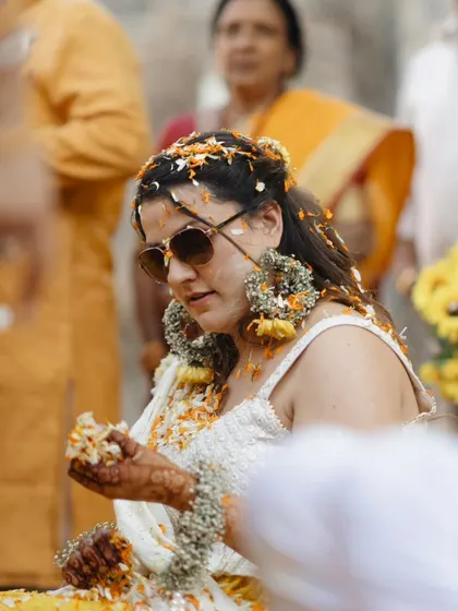 The bride, adorned with floral jewelry, takes a moment during her Haldi ceremony. The sunglasses add a cool, modern twist to the traditional event.
