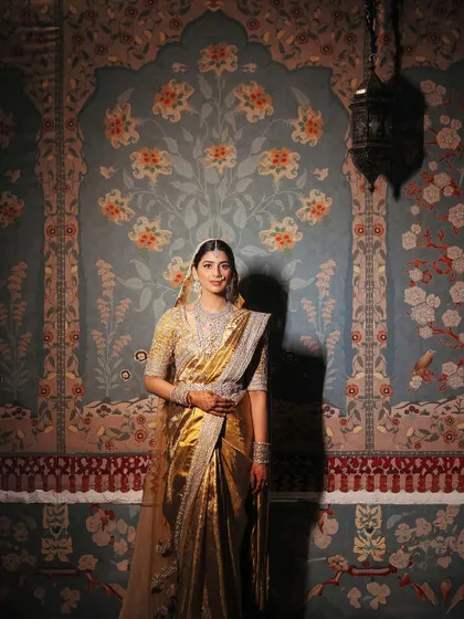A bride stands like a portrait against a wall with traditional Indian frescoes. Her golden saree and classic jewelry create a look of timeless elegance for her welcome dinner.