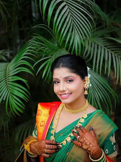The bride looks directly at the camera, her hands gently clasped. The lush green foliage in the background complements her beautiful green saree and traditional jewelry.