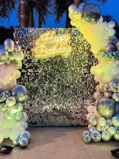 An outdoor disco party setup at dusk. The silver shimmer wall and balloon arch are lit up by the neon sign and colored lights, creating a magical effect.
