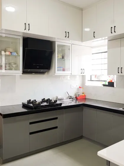 A classic two-tone combination of glossy grey base cabinets and white wall cabinets. This L-shaped modular kitchen is designed for functionality, with glass-fronted display cabinets and sleek black handles adding a modern touch.