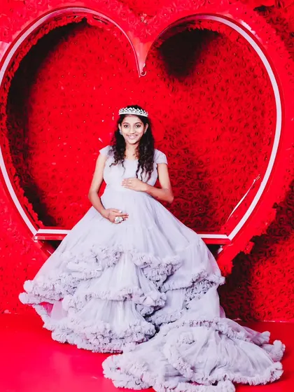A full-length shot of an expectant mother in a beautiful light purple ruffled gown, posing in front of a large red heart.