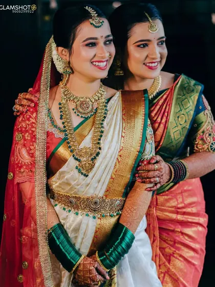 The pure joy of a bride with her sister. She is wearing a beautiful temple jewellery set with green stones that pop against her white and gold silk saree.