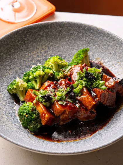 A dish of braised pork belly with broccoli. The lighting is carefully controlled to highlight the rich glaze on the meat and the vibrant green of the vegetable.