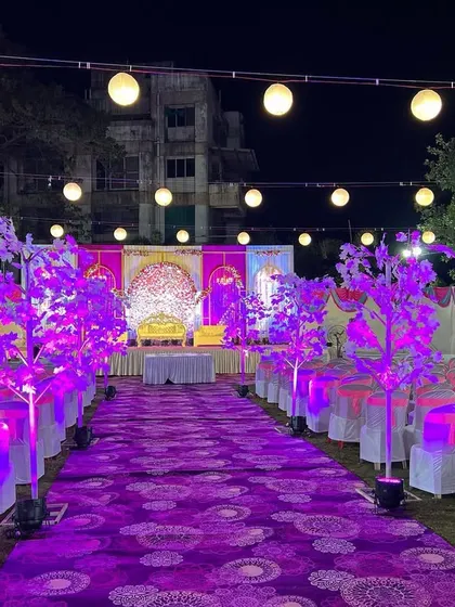 The full view of an outdoor wedding reception area with a purple and pink theme. The long aisle is lined with illuminated decorative trees, leading the eye directly to the grand stage.