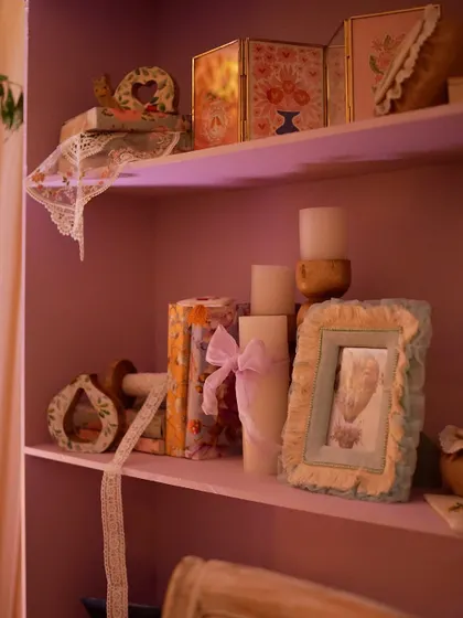 A shelf painted in a soft lavender hue, styled with candles, books, and framed pictures.