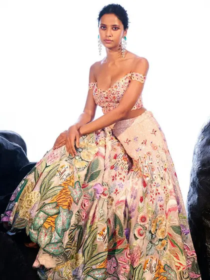 For a modern bride, this beige lehenga features a unique off-shoulder blouse and a skirt adorned with tropical floral and leaf motifs. It's a fresh and contemporary take on bridal wear.