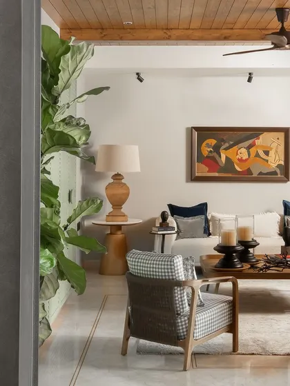 A wider view of the living room, showing how the large plant and architectural details frame the space. The MF Husain painting remains a powerful focal point, balanced by the room's earthy and neutral tones.