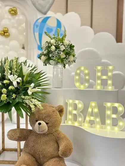 A close-up of the 'Oh Baby' marquee letters and teddy bear detail. The warm lights and lush floral arrangements add a touch of elegance and warmth to the overall setup.