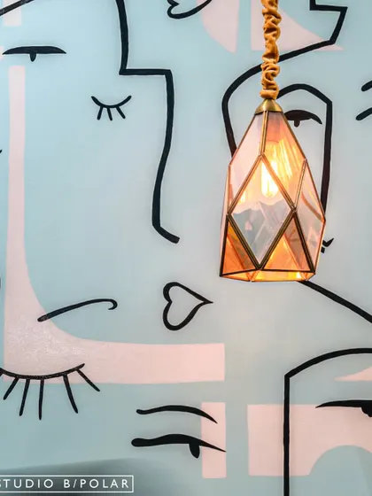 A close-up of a custom wallpaper with abstract face line art, paired with an elegant geometric pendant light. This detail shot highlights our focus on combining artistic elements and unique lighting to enrich a space.