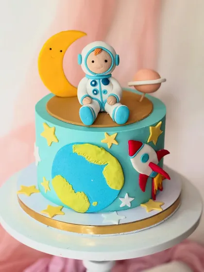 A cute outer space cake with a friendly astronaut. This design features a fondant astronaut, moon, planet, and rocket ship on a space-themed buttercream cake.