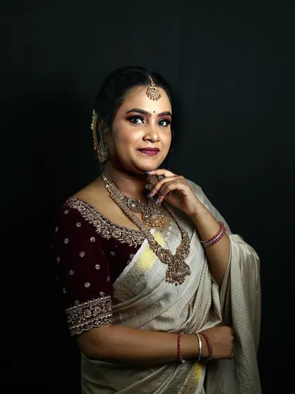 A studio portrait showcasing a bridal look with a modern twist. The makeup features a dramatic eye, while the off-white saree and maroon velvet blouse create a unique color combination.