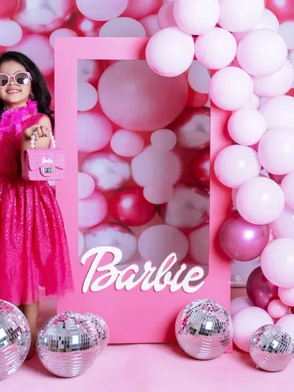 A vibrant 'Barbie' themed photoshoot for a stylish toddler. She poses confidently in a pink frame against a wall of pink balloons and disco balls, complete with a feather boa and sunglasses.