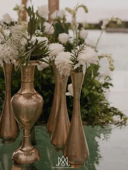 A close-up of elegant brass vases holding simple white floral arrangements, placed on the glass floor of the water mandap.