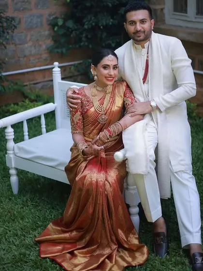 A full portrait of the couple, showcasing the groom's clean and modern white kurta-pajama set. The simplicity of his outfit allows the bride's traditional attire to take center stage.