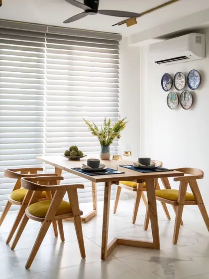 This view highlights the clean, minimalist aesthetic of the dining area. The simple wooden table and chairs are set against a bright white wall, with natural light filtering through the blinds for a fresh, airy feel.