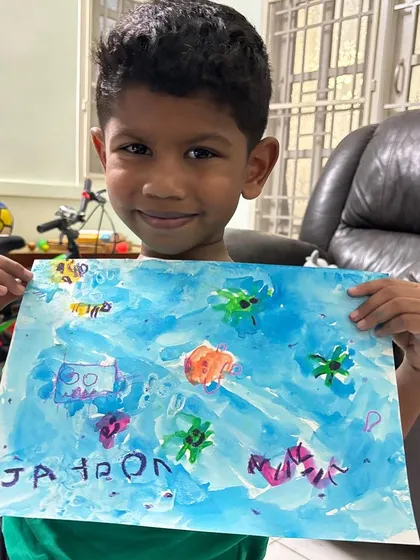 A proud young artist from our online class holding up his underwater scene. Our virtual sessions are filled with stories and fun themes to keep children engaged and excited to learn.