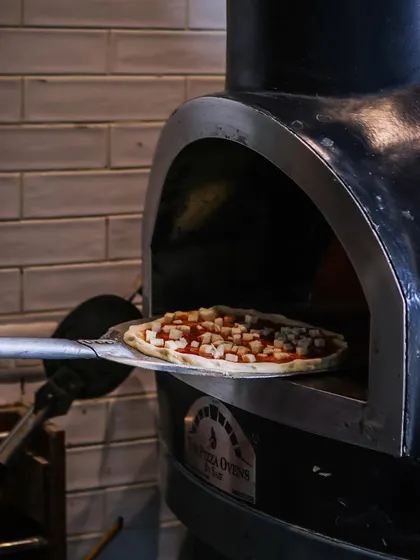 Pizza in progress. A pie topped with cheese cubes is ready to slide into the fiery oven. This is the moment before the transformation begins.