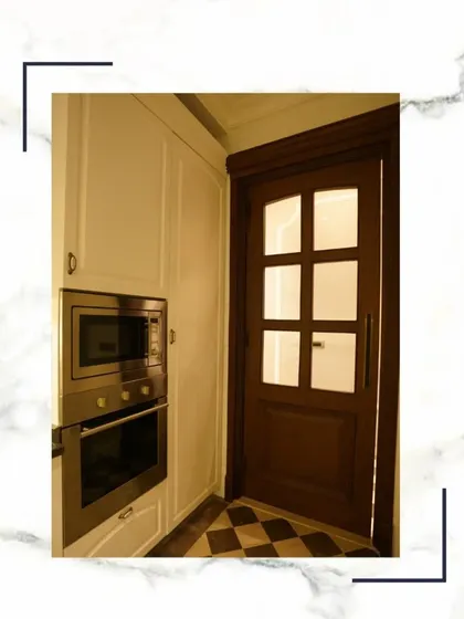 A modern kitchen with built-in microwave and oven, seamlessly integrated into white cabinetry. The classic wooden door and checkerboard floor add a touch of timeless style.