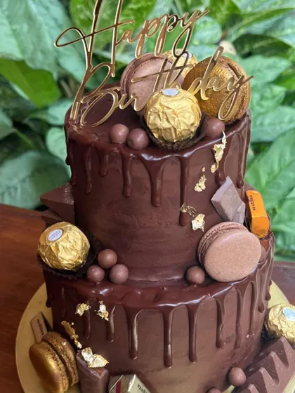 A decadent two-tier chocolate drip cake topped with Ferrero Rocher, macarons, and gold leaf. I also offer designer cakes to complement my gifting services.