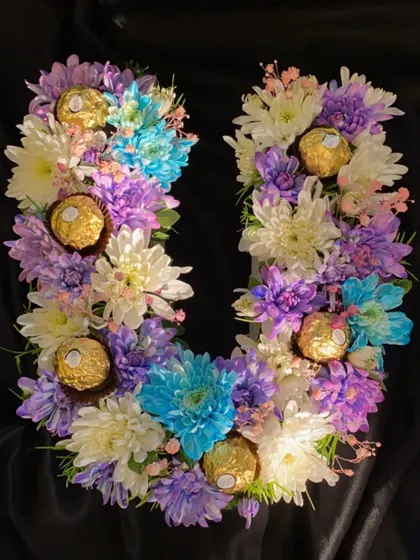 A full view of the custom 'U' monogram bouquet against a dark backdrop, making the bright colors of the chrysanthemums and the gold of the Ferrero Rocher pop.