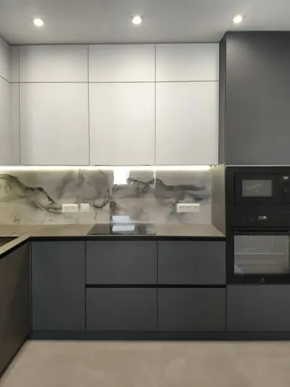A minimalist L-shaped kitchen combining dark grey base cabinets with white upper units. The unique marbled backsplash and under-cabinet lighting create a focal point, while the integrated oven and microwave unit ensures a streamlined appearance.