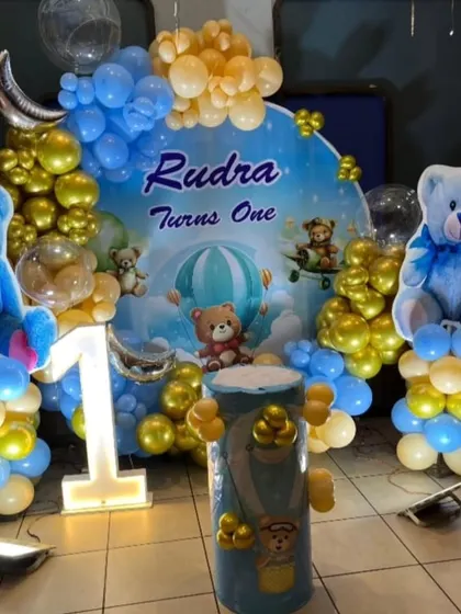 A charming teddy bear themed first birthday setup. The decor features a large backdrop, big blue teddy bear figures, a light-up number "1", and balloons in shades of blue and gold.