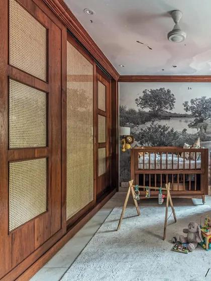 This shot shows the scale of the room, with the large, custom-built wooden and cane wardrobe on the left and the cozy crib and play area on the right. It’s a space that is both grand and intimate.