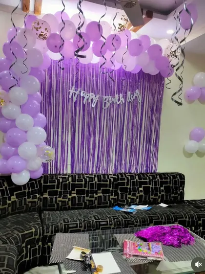 A simple birthday decoration in a living room with a purple shimmer curtain and a purple and white balloon arch.
